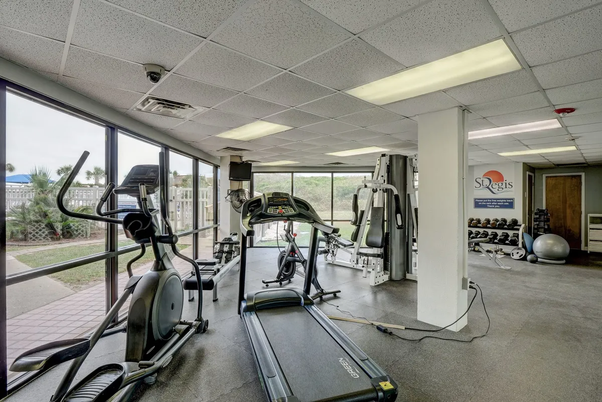 A bright and motivational fitness center featuring a treadmill, elliptical machine, weight lifting equipment, and large windows letting in natural light. The setup encourages exercise and healthy routines.
