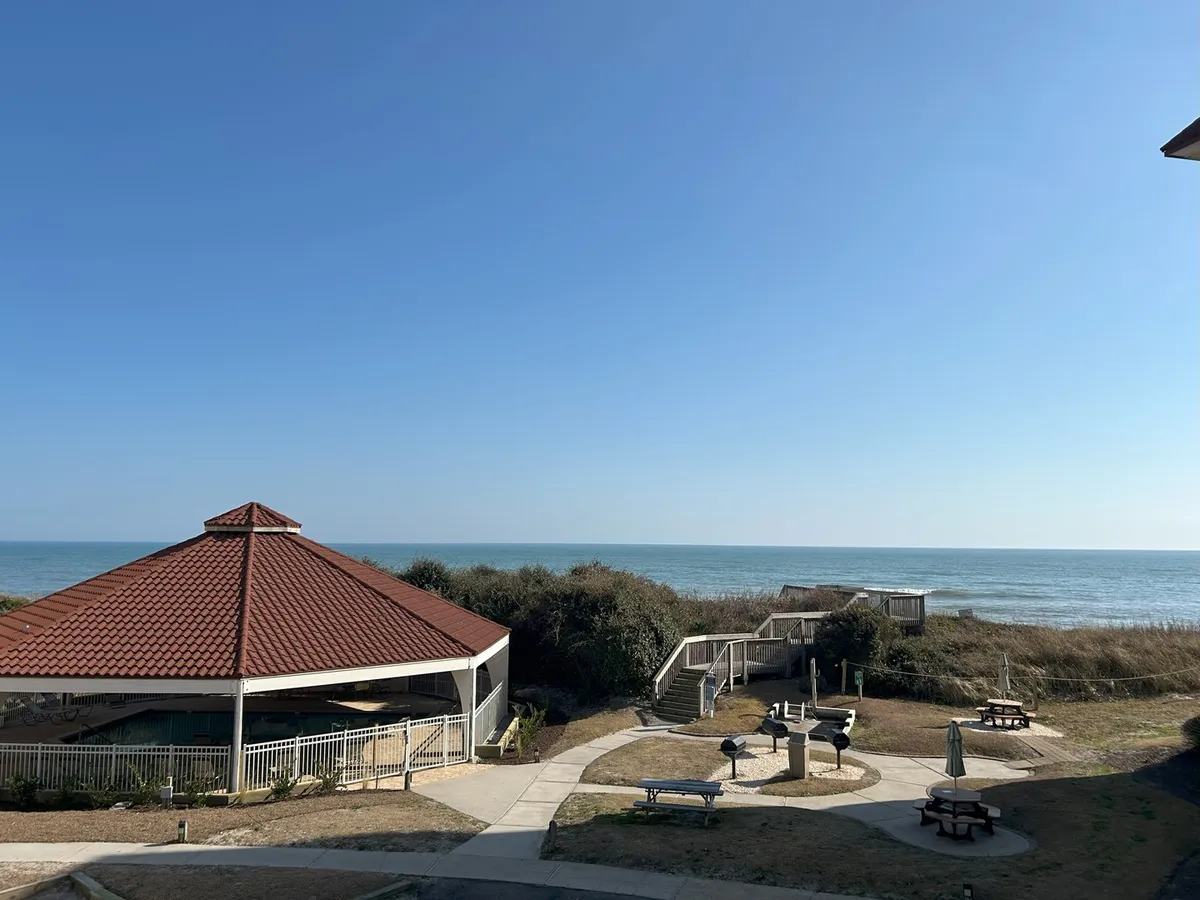 A serene outdoor area with a gazebo, picnic tables, and pathways all leading towards a stunning ocean backdrop. The space is designed for gatherings, barbecues, and leisurely strolls, with the beach and water visible just beyond.