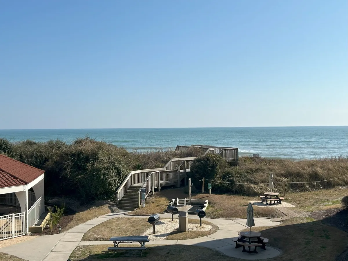 This exterior image captures an inviting outdoor area with comfortable seating, barbecue grills, and a direct view of the ocean. A staircase leads down toward the sandy beach, suggesting easy access for morning walks or quick breaks by the waves. The scene blends relaxation with activity, featuring spaces for picnicking, barbecuing, or simply taking in the coastal atmosphere.