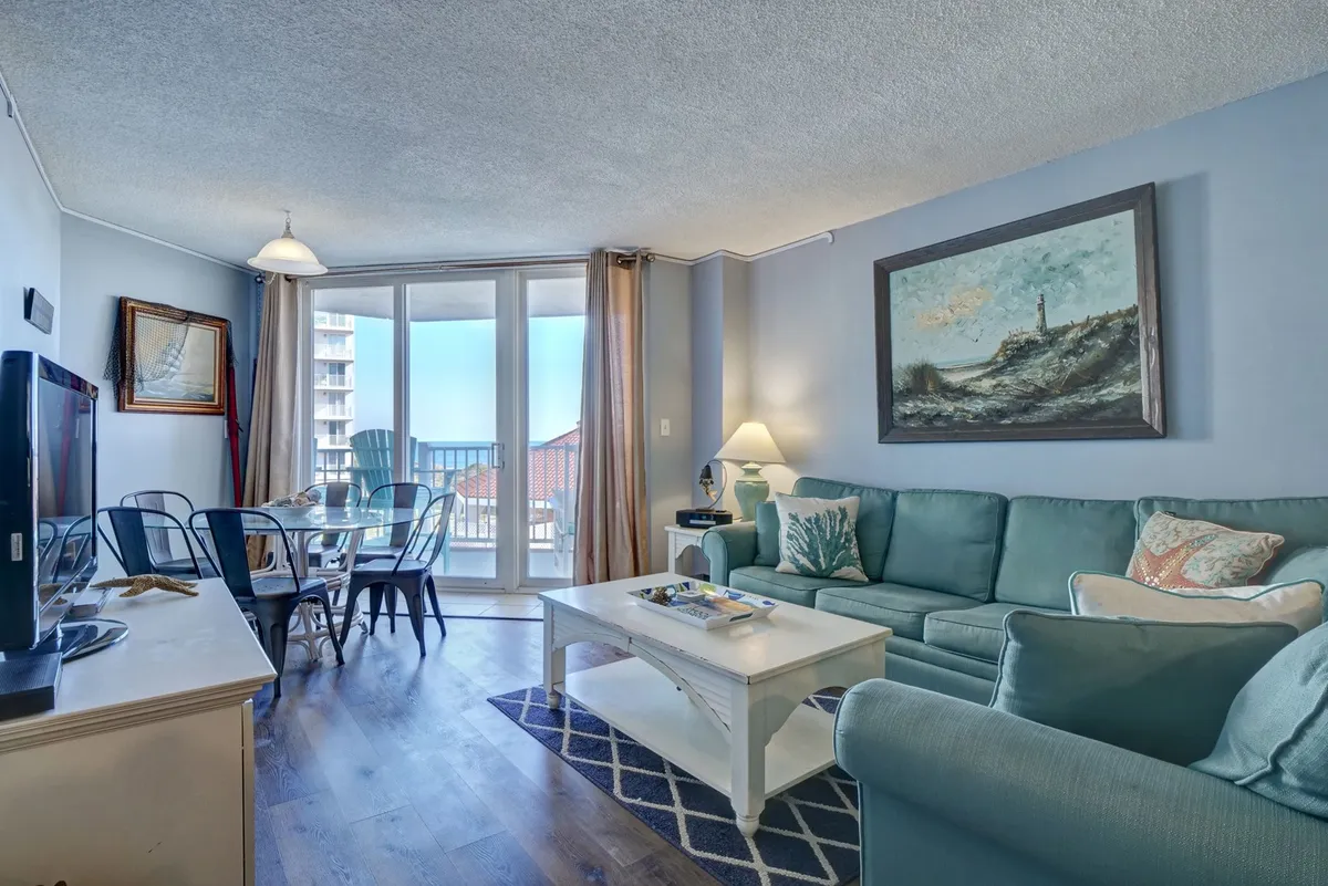A cozy view of the living room featuring a comfortable sofa, dining table, large wall art, and balcony access. The space exudes warmth and casual elegance, emphasizing areas where guests can gather for meals, conversation, or movie nights.