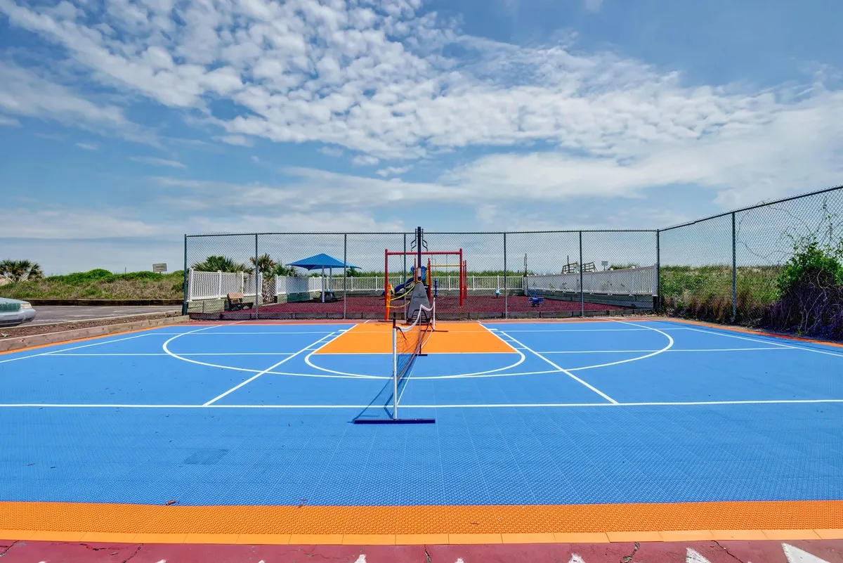 A striking blue sports court with orange accents, complete with a tennis net and adjacent playground equipment, set in an active outdoor environment. The space is designed for both basketball and tennis, promoting active fun for all ages.
