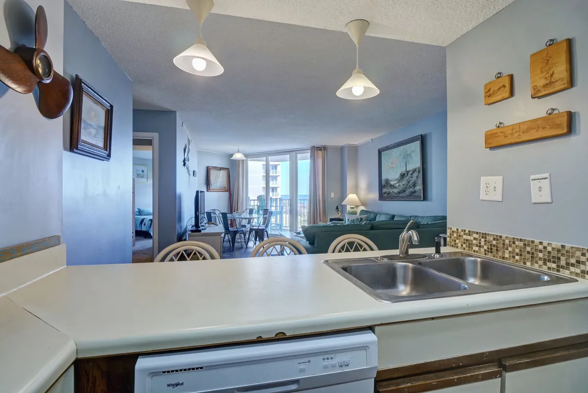 A bright, open-concept living area seamlessly flowing into the dining space and kitchen. The image frames the spacious kitchen counter, dining table, inviting living room with a large sectional, and a balcony view with the ocean beyond, all bathed in natural light from floor-to-ceiling windows.