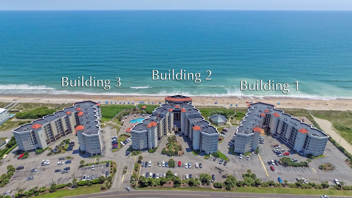 An elevated view shows three resort buildings framing a large beachfront swimming pool with ample lounge seating. The pool is just steps away from the sandy beach beyond, offering a seamless connection between swimming, sunbathing, and beach walks. Families gather around the pool area, and the proximity to the ocean is clear.