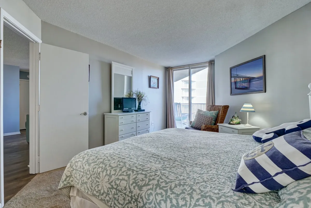 A cozy bedroom featuring a bed with a patterned cover, a sliding glass door that likely leads to a private balcony, a mirror and dresser, and an armchair with soft cushions. Sunlight streams into the room, and there’s a framed picture on the wall, creating a calm, inviting space perfect for unwinding.