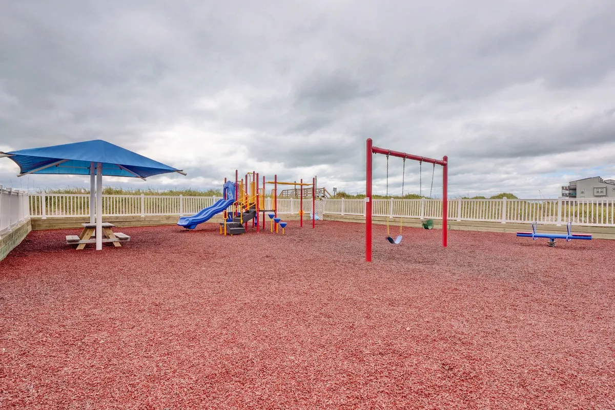 A vibrant playground with colorful slides, swings, and a picnic table under a blue umbrella. Children are playing happily, and the area is bathed in cheerful sunlight.