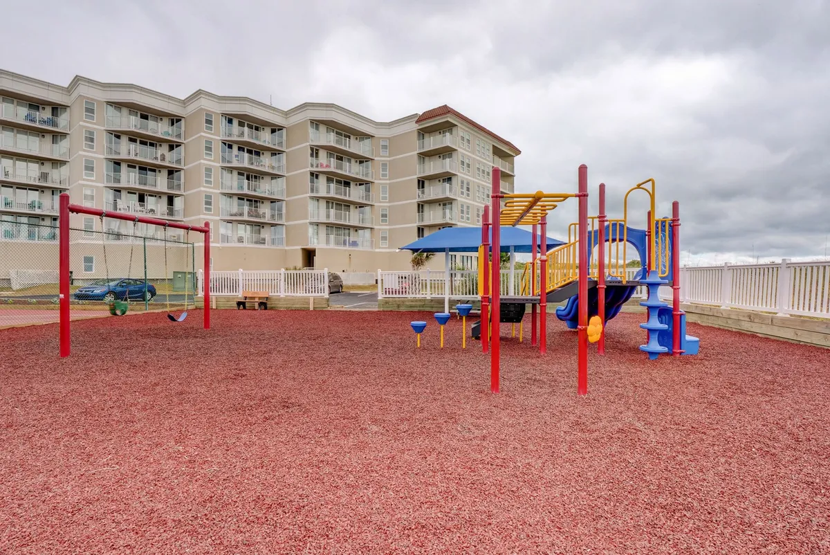 A vibrant playground with colorful equipment and swings sits outdoors, surrounded by open space for children to run and play. The mood is lively and joyful, with kids actively enjoying the playground, making it clear that the property caters to families with young children.