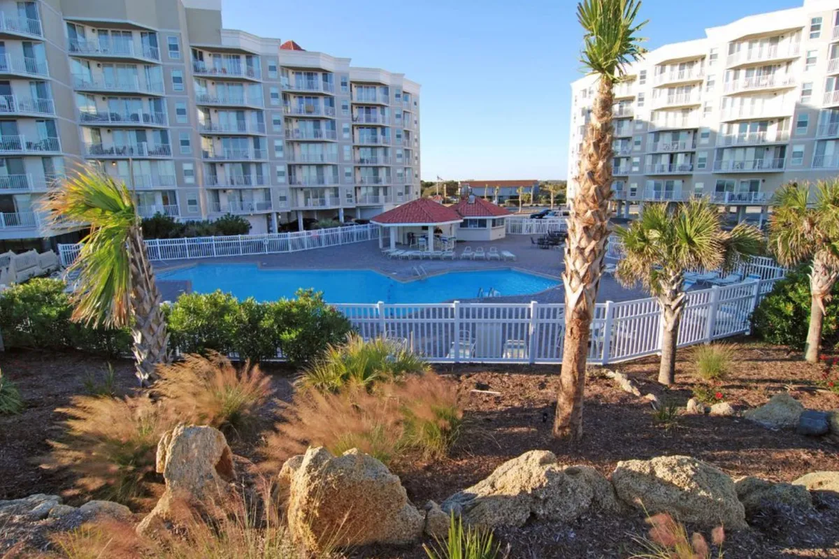 A sparkling swimming pool surrounded by palm trees and an expansive patio area, with the resort building visible in the background. The inviting setting is perfect for swimming, sunbathing, and socializing outdoors.