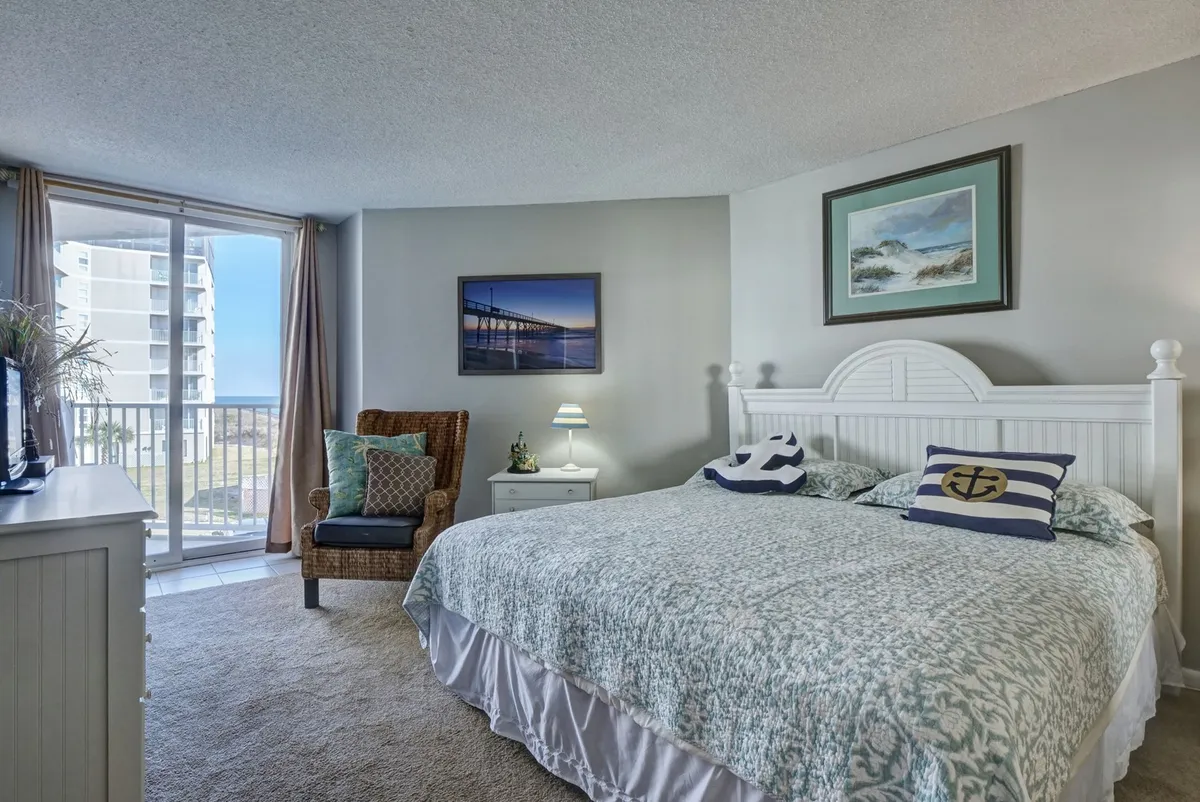 A serene bedroom with a queen-sized bed dressed in patterned bedding, a cozy armchair beside large windows that flood the room with natural light, and decorative artwork on the walls. The room exudes calm and relaxation, with views framed by the windows, hinting at the proximity to the shore.