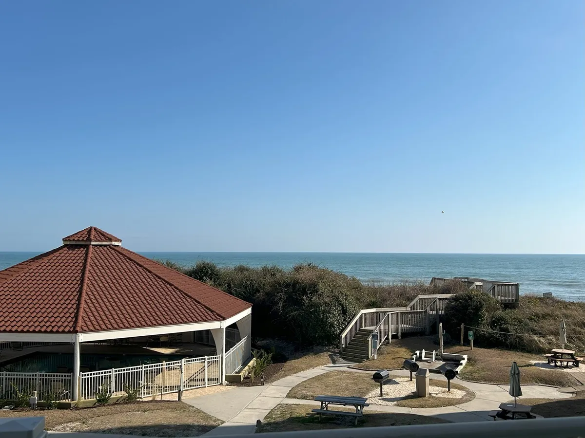 The image showcases the inviting outdoor amenities of the St Regis condo complex at North Topsail Beach. A charming gazebo and grilling area are set along a manicured pathway, leading guests toward sweeping ocean views. The scene suggests tranquil moments spent picnicking, barbecuing with family or friends, and strolling toward the sand, all framed by the backdrop of the Atlantic and the promise of stunning sunsets.