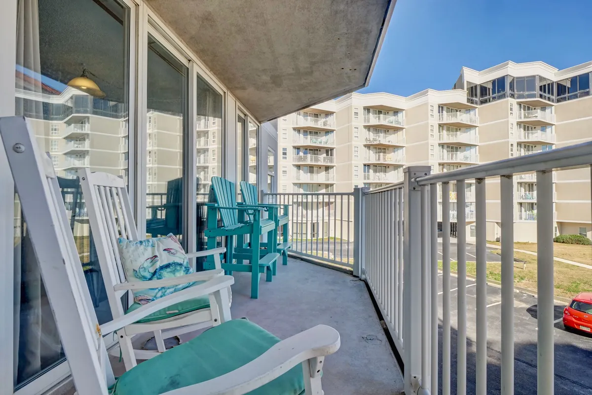 An exterior shot of a private balcony furnished with both rocking chairs and colorful Adirondack chairs, overlooking the property and offering a tranquil setting for socializing, morning coffee, or sunset contemplation. The view extends over the parking area toward the horizon, providing a sense of openness and peaceful retreat.